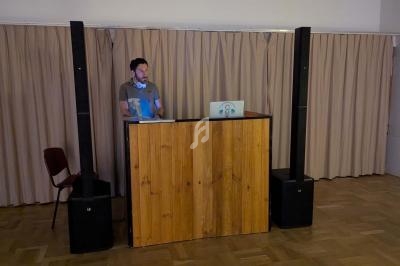 Un homme debout derrière un pupitre en bois avec un ordinateur portable, dans une salle avec rideaux beiges.