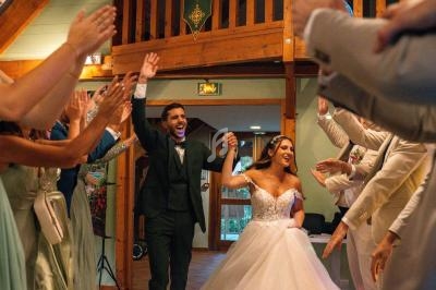 Un couple de mariés souriants entre dans une salle en bois, acclamé par des invités levant les bras.