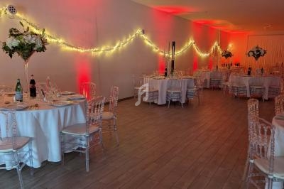 Salle de réception décorée avec des tables rondes, nappes blanches, chaises transparentes et guirlandes lumineuses.