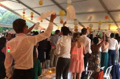 Des invités debout sous une tente festive agitent des serviettes blanches lors d'une célébration animée.