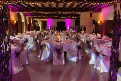 Salle de réception décorée avec des tables rondes, nappes blanches, chaises ornées de rubans violets et éclairage tamisé.
