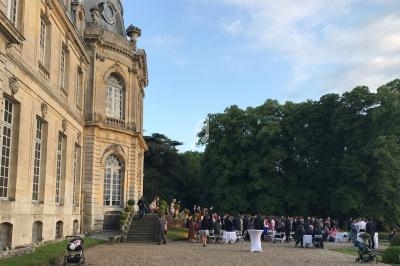 Groupe de personnes rassemblées devant un château ancien, avec des arbres en arrière-plan et un ciel partiellement dégagé.