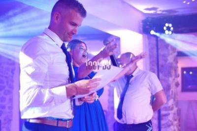 Un homme lit un document pendant qu'une femme chante au micro dans une salle éclairée par des lumières colorées.