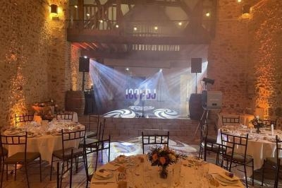 Salle en pierre avec tables dressées pour un repas, éclairage chaleureux et scène équipée de projecteurs.