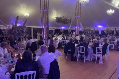 Invités assis à des tables rondes dans une grande tente éclairée de lumières violettes, décorée de fleurs suspendues.