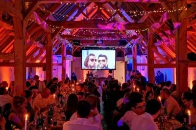 Salle de réception en bois décorée de guirlandes lumineuses, avec des invités assis et un écran projetant une image.