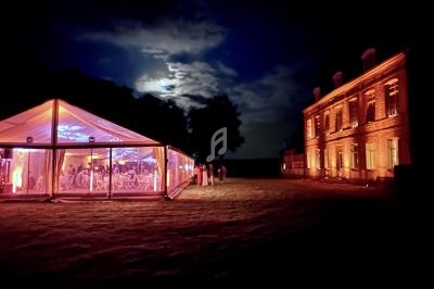 Tente éclairée et bâtiment illuminé dans un jardin de nuit sous un ciel partiellement nuageux.