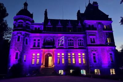 Façade d'un château illuminée en violet à la tombée de la nuit, avec des fenêtres éclairées et un ciel sombre.