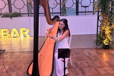 Une femme en robe blanche joue de la harpe dans une salle décorée de plantes et de lettres lumineuses ’BAR’.