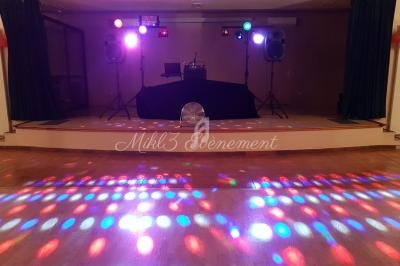 Scène de soirée avec jeux de lumières colorés, platine DJ et enceintes sur une estrade dans une salle.