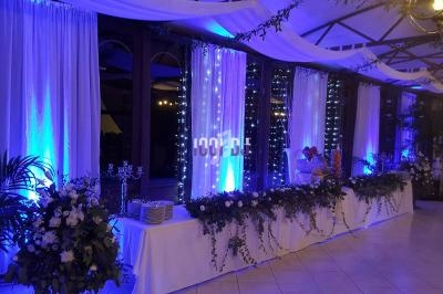 Décoration de salle de réception avec rideaux blancs, éclairage bleu, guirlandes lumineuses et tables ornées de fleurs.