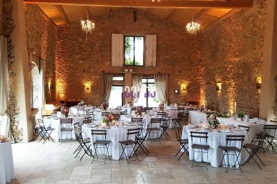 Salle de réception en pierre avec tables rondes dressées, chaises en bois et lumière naturelle entrant par de grandes…