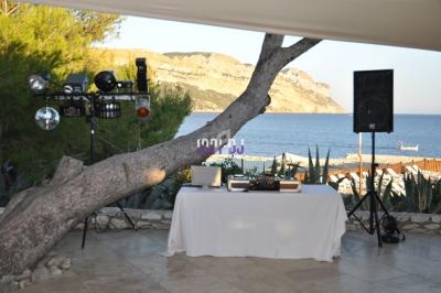 Table de mixage et éclairages installés en extérieur près d'un arbre, avec vue sur la mer et des collines en arrière-plan.