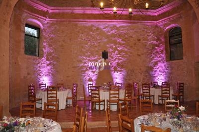 Salle en pierre avec tables dressées pour un événement, éclairée par des lumières roses et un lustre suspendu.