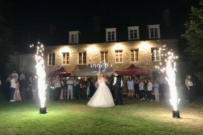 Un couple danse en tenue de mariage devant des invités, avec des étincelles et une maison illuminée en arrière-plan.