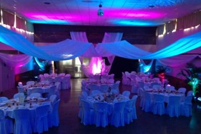 Salle de réception décorée avec des tables rondes, nappes blanches, éclairages colorés et voilages suspendus.