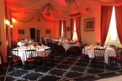Salle de réception décorée avec des tables dressées, nappes blanches, chaises noires et éclairage rouge chaleureux.