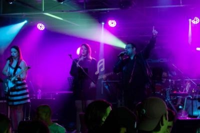 Groupe de musiciens et chanteurs sur scène, éclairés par des lumières violettes, devant un public.