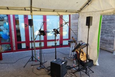 Équipement de sonorisation avec micros, guitare et amplificateur installé sous une tente devant un bâtiment vitré.