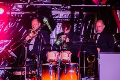 Musiciens jouant du trombone, de la trompette et du saxophone sur une scène éclairée, avec des percussions au premier plan.