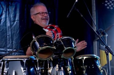 Un musicien joue des congas sur scène, tenant un tambourin, sous un éclairage de concert.