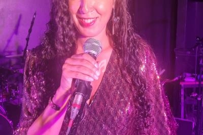 Femme souriante en robe à paillettes violettes tenant un micro sur une scène éclairée de lumières roses.