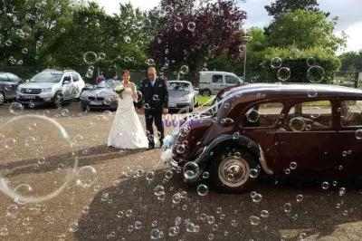 Un couple de mariés marche près d'une voiture ancienne, entouré de bulles, dans un cadre extérieur verdoyant.