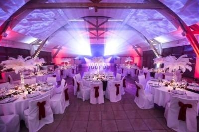 Salle de réception décorée avec des tables rondes, nappes blanches, chaises ornées de nœuds rouges et éclairage coloré.