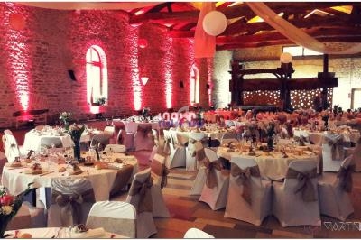 Salle de réception décorée pour un mariage, avec tables rondes dressées, chaises ornées de nœuds et éclairage chaleureux.