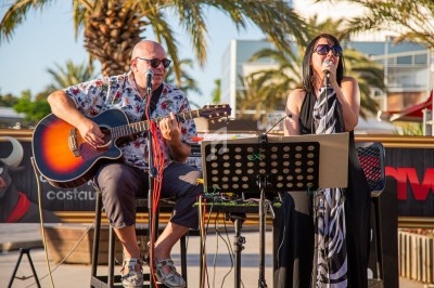 Un homme joue de la guitare et une femme chante sur une scène extérieure, avec des palmiers en arrière-plan.