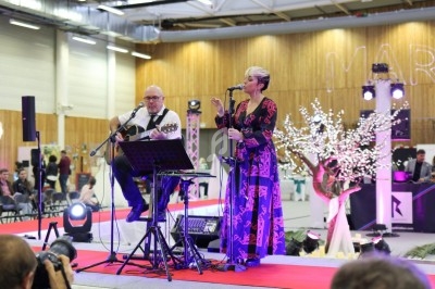 Un duo musical se produit sur une scène intérieure avec guitare et chant, devant un public assis.