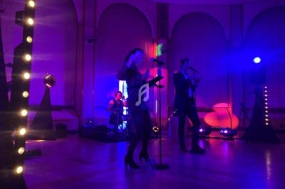 Un groupe joue sur scène dans une salle éclairée par des lumières colorées, avec un batteur, une chanteuse et un…