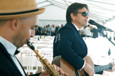 Un chanteur joue de la guitare et chante au micro tandis qu'un musicien joue du saxophone sous une tente blanche.