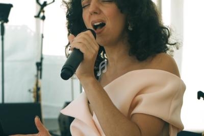 Femme chantant avec émotion dans un micro, vêtue d'une robe claire, devant un fond lumineux et des instruments.