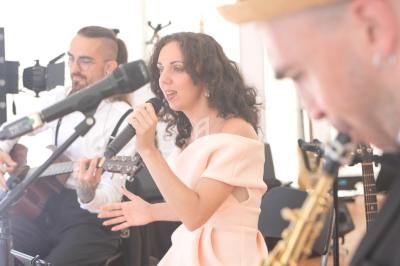 Une chanteuse interprète un morceau au micro, accompagnée d'un guitariste et d'un saxophoniste en arrière-plan.