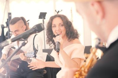 Une chanteuse souriante interprète une chanson, accompagnée d'un guitariste et d'un saxophoniste en arrière-plan.