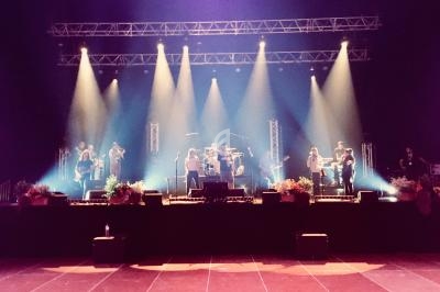 Groupe de musiciens sur scène illuminée par des projecteurs, avec des fleurs disposées au premier plan.
