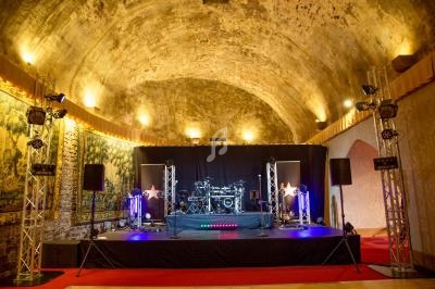 Scène de concert éclairée sous une voûte en pierre, entourée de tapisseries et de projecteurs.