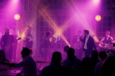 Groupe de musiciens et chanteurs sur scène avec éclairage violet, devant un public dansant dans une salle.