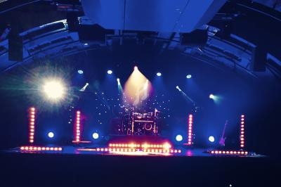 Scène de concert éclairée par des projecteurs colorés, avec une batterie et une guitare visibles au centre.