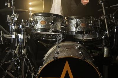 Un batteur en costume joue sur une batterie argentée, éclairé par des projecteurs en arrière-plan.