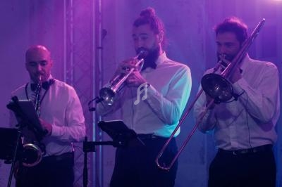 Trois musiciens en chemise blanche jouent du saxophone, de la trompette et du trombone sur une scène éclairée en violet.