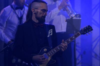Un guitariste en costume joue sur scène, entouré de musiciens en arrière-plan sous un éclairage bleu.