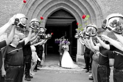 Un couple de mariés s'embrasse sous une haie d'honneur formée par des pompiers tenant des roses rouges.