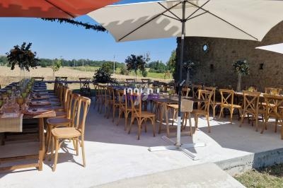 Tables en bois dressées pour un repas en extérieur, sous des parasols, avec vue sur un champ et des décorations florales.