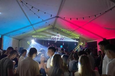 Foule rassemblée sous une tente éclairée par des lumières colorées, avec un DJ sur scène en arrière-plan.