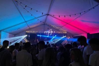 Foule rassemblée sous une tente éclairée par des lumières colorées, avec une scène et un DJ en arrière-plan.