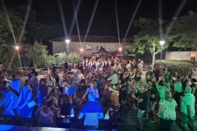 Groupe de personnes dansant en plein air la nuit, éclairées par des lumières, avec des arbres et un bâtiment en arrière-plan.