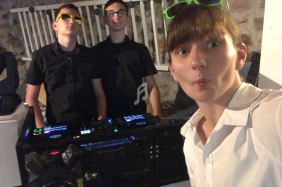 Trois jeunes posent devant une table de mixage éclairée, deux portant des lunettes de soleil colorées.