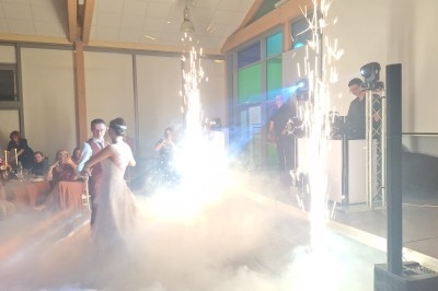Un couple danse dans une salle éclairée, entouré de fumée et d'effets pyrotechniques, devant des invités assis.
